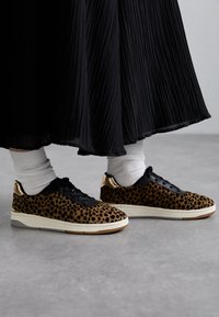Leopardmønstrede sneakers med sort tekstiloverdel, hvid sål og guldfarvede detaljer på hælen. Båret med hvide sokker og en sort plisseret nederdel.