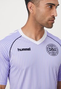 Uomo con maglia sportiva a righe lavanda e bianche con il logo "hummel" e l'emblema della Danish Boldspil-Union (DBU), che guarda verso destra.
