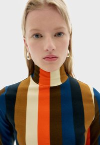 Fitted turtleneck top featuring bold vertical stripes in blue, orange, brown, and cream, made from smooth, stretchy fabric.