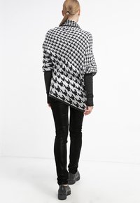 Czarnobiały ponczo w wzór pepitki, z drapowanym krojem, w połączeniu z czarnymi dopasowanymi spodniami i ciemnymi butami.