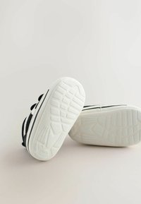 Black canvas sneakers with white rubber soles, featuring a textured tread pattern and black lace accents. View from the back.