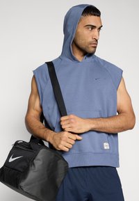 Sweat à capuche sans manches en tissu bleu discret avec une poche kangourou devant ; accompagné d'un sac de sport noir avec un logo Nike blanc.