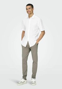 Camisa blanca de manga corta y botones, pantalones beige de corte ajustado y zapatillas blancas con detalles grises. El modelo está de pie con las manos en los bolsillos.