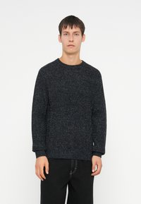 Joven de pie, de frente, con un suéter de punto texturizado oscuro y pantalones negros, contra un fondo blanco.