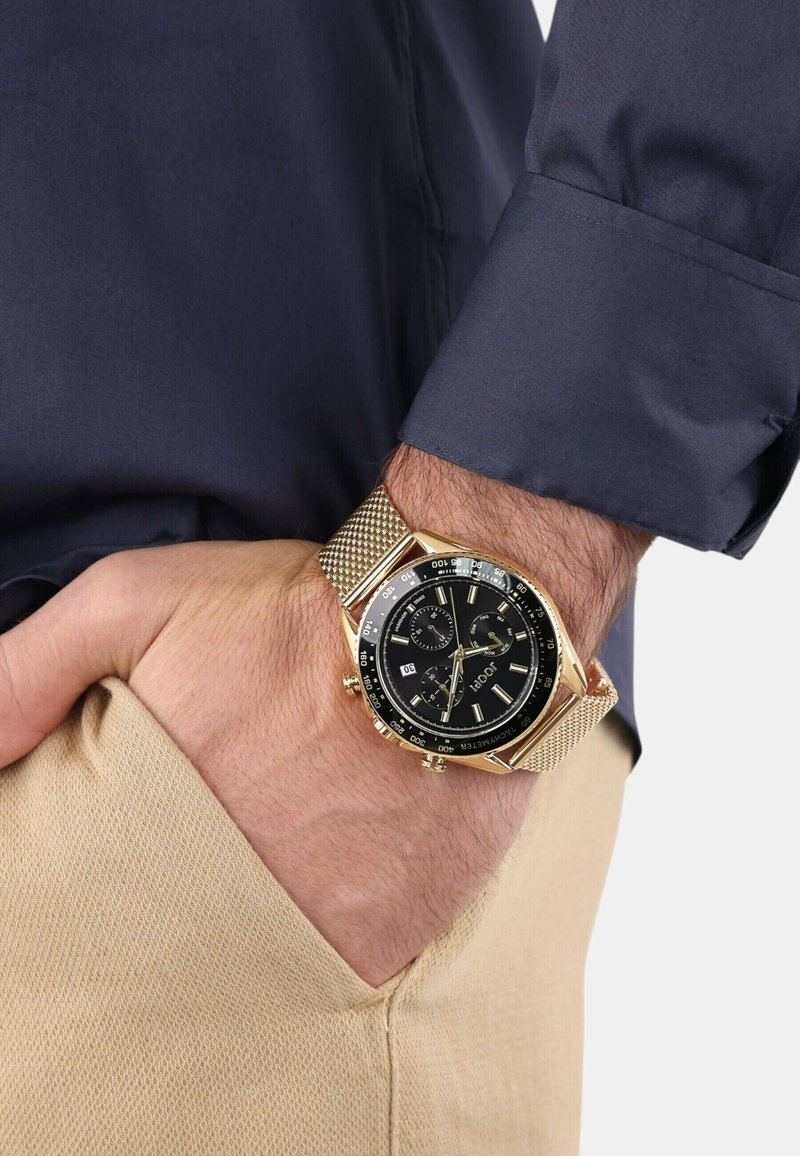 Herrenarmbanduhr mit schwarzem Zifferblatt, Gehäuse in Goldfarbe und Mesh-Armband. Zu den Merkmalen gehören Subzifferblätter und eine Datumsanzeige. Getragen auf einer Hand in einem marineblauen Ärmel.