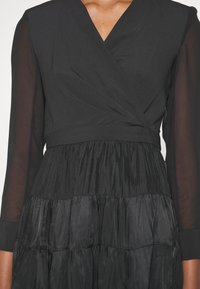 Robe noire avec manches longues transparentes, corsage croisé en V et jupe à volants froncée sur une personne.