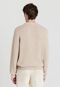 Mann in beigem strukturiertem Strickpullover und cremefarbenen Hosen, von hinten vor einem schlichten hellen Hintergrund gezeigt.