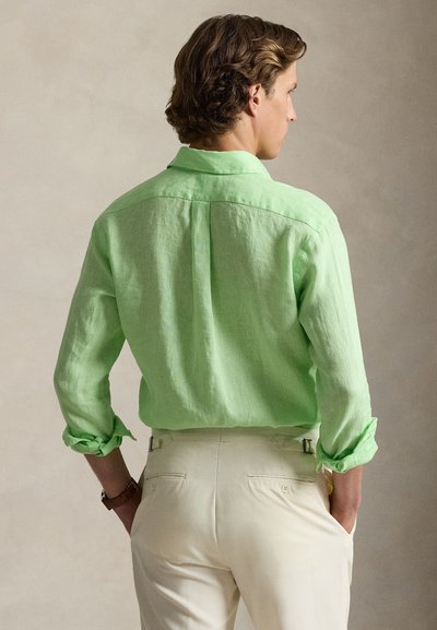 Ανοιχτό πράσινο πουκάμισο από λινό με τα μανίκια γυρισμένα προς τα πάνω, απεικονιζόμενο από πίσω. Συνδυάζεται με κρεμ παντελόνι, με ρυθμιζόμενα πλαϊνά λουράκια.