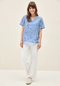 Maglietta blu a maniche corte con scollo a V e motivo a foglia bianca, abbinata a pantaloni bianchi a gamba larga e sneakers beige su uno sfondo neutro.