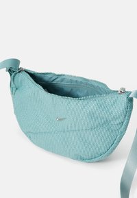 Turquoise fabric bag with a textured surface, silver zipper, and small silver logo. Features a slim, curved design and adjustable strap.