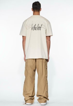 Cream-colored oversized t-shirt featuring graphic text on the back, paired with loose-fitting khaki cargo pants and white sneakers.