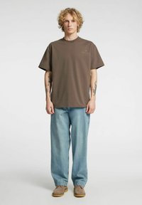 Rjava bombažna majica s kratkimi rokavi, kombinirana s svetlo modrimi širokimi kavbojkami in sivimi semiš čevlji. Preprost dizajn, brez vidnih vzorcev.