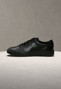 Baskets basses en cuir noir avec lacets noirs et marquage en relief subtil sur le côté, présentées sur une surface neutre.