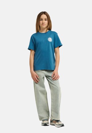 Jeune personne debout portant un t-shirt graphique bleu, un jean large délavé clair et des baskets multicolores sur fond blanc.