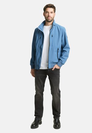 Uomo in piedi che indossa una giacca azzurra con zip sopra una maglietta bianca, jeans scuri e stivali neri, con una mano in tasca.