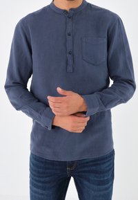 Uomo che indossa una maglietta henley a maniche lunghe di colore blu navy e jeans blu, che aggiusta una manica con entrambe le mani su uno sfondo bianco.
