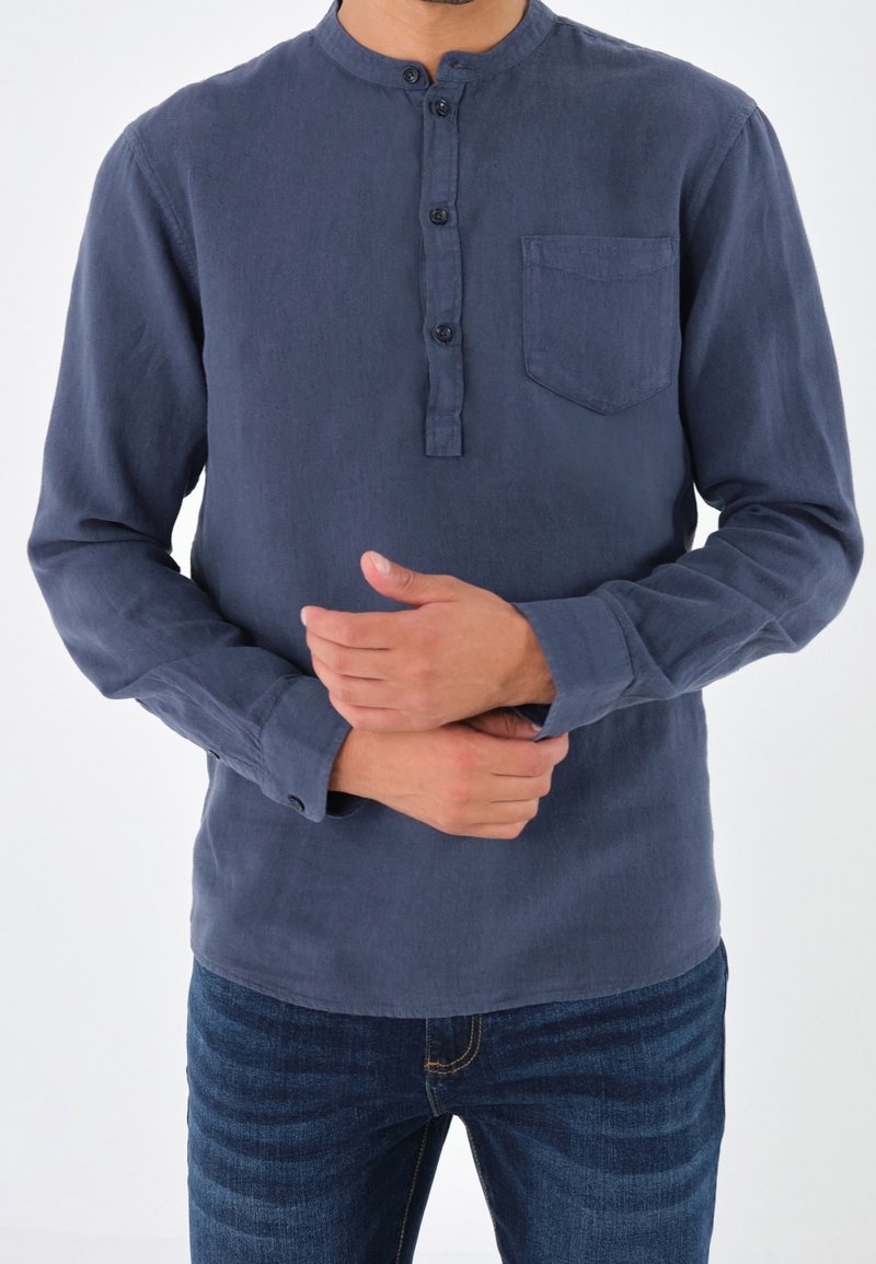 Uomo che indossa una maglietta henley a maniche lunghe di colore blu navy e jeans blu, che aggiusta una manica con entrambe le mani su uno sfondo bianco.