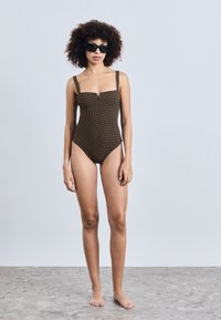 Brunmønstret badedrakt med tykke stropper, som har firkantet utringning og subtil V-kutt-detalj. Modellen har på seg svarte solbriller og er barføtt.