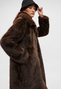 Hnedý kabát z umelého kožušina s vysokým golierom a dlhými rukávmi, ktorý má mäkký, textúrovaný povrch. Nosí sa s čiernou pletenou čiapkou.