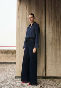 Chemise en denim avec col et poche, associée à un jean bleu foncé à coupe large. Présente une coupe décontractée et une couture visible. Des chaussures rouges vives complètent le look.