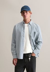 Jasnoniebieska koszula na guziki z kieszenią na piersi, noszona na białej koszulce; w połączeniu z czarnymi dżinsami i ciemnoniebieską czapką.
