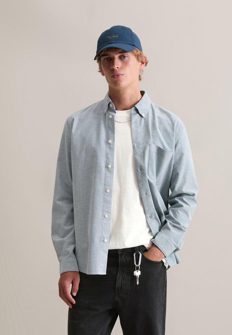 Jasnoniebieska koszula na guziki z kieszenią na piersi, noszona na białej koszulce; w połączeniu z czarnymi dżinsami i ciemnoniebieską czapką.