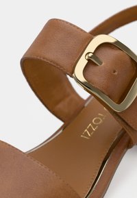 Καφέ συνθετικό δερμάτινο σανδάλι με φαρδιά λουράκι στον αστράγαλο, διακοσμημένο με μεγάλη χρυσή αγκράφα και απαλό εσωτερικό επένδυση. Μινιμαλιστικός σχεδιασμός.