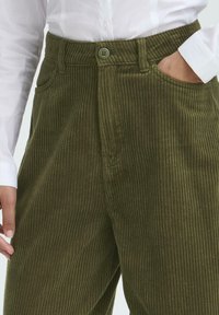 Pantalone od maslinaste korduroj tkanine s vertikalnom rebrastom teksturom, s kopčanjem na dugme i bočnim džepovima. Klasičan dizajn za svakodnevno nošenje.