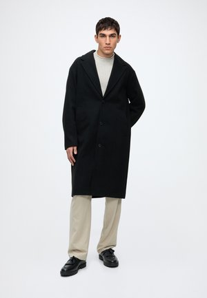 Homme portant un long manteau noir, un pull clair, un pantalon beige et des chaussures noires, debout sur un fond blanc uni.