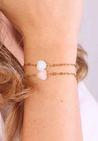 Gouden kettingarmband met twee hartvormige bedels, één lichtblauw en één bleekroze. Glad oppervlak, elegant ontwerp, gedragen om een pols.