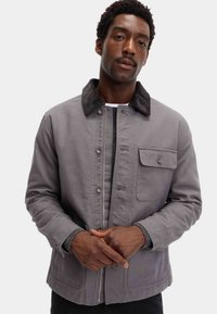 Grå stofjakke med en kordfløjlskrave, knapper foran og en brystlomme. Har et struktureret design og kontrasterende teksturer.