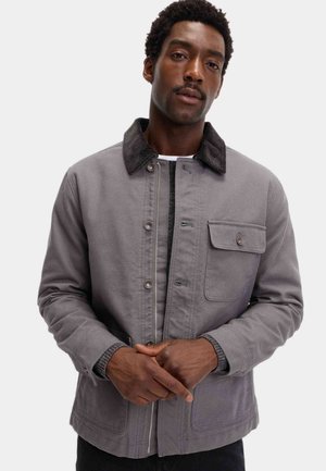 Grijze stoffen jas met een corduroy kraag, knopen aan de voorkant en een borstzak. Heeft een gestructureerd ontwerp en contrasterende texturen.