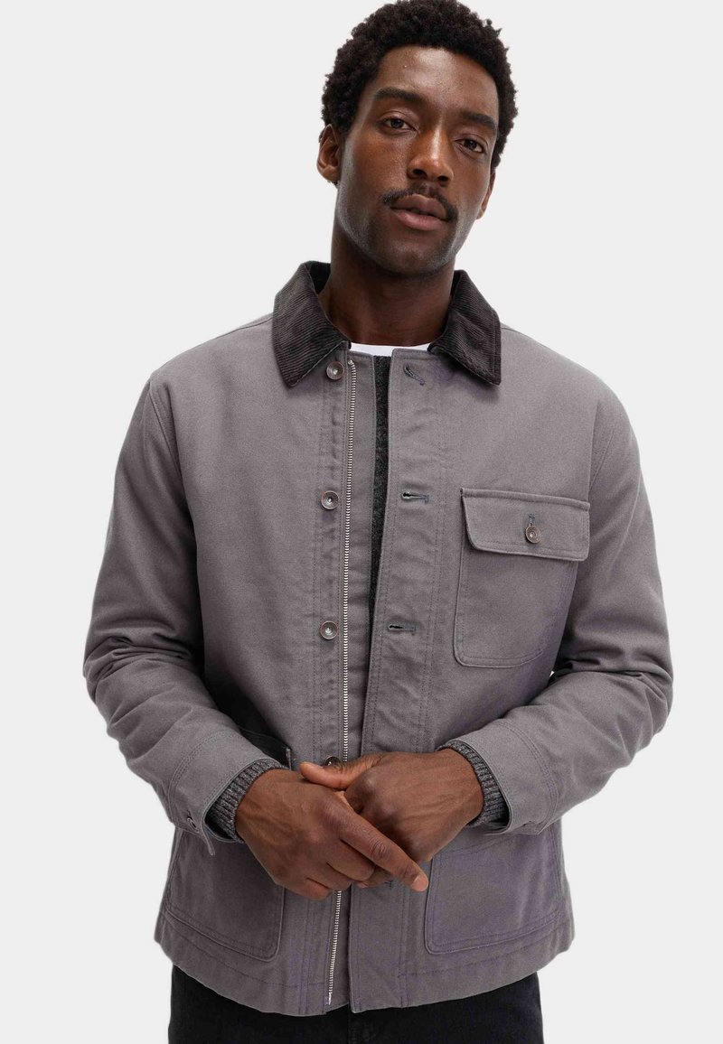Grå stofjakke med en kordfløjlskrave, knapper foran og en brystlomme. Har et struktureret design og kontrasterende teksturer.