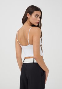 Camisola bianca a coste, corta, con spalline sottili, abbinata a pantaloni neri a vita alta con dettagli a contrasto sulla cintura.