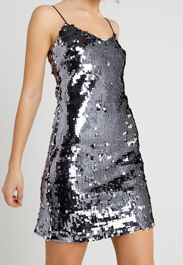 Femme portant une mini-robe argentée sans manches à sequins avec fines bretelles spaghetti sur un fond clair uni.