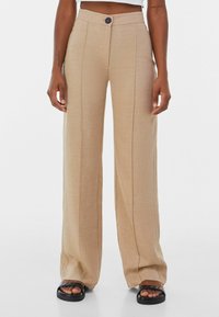 Beige weit geschnittene Hose mit glatter Textur, Bügelfalte und einem einzelnen Knopfverschluss. Tragen mit schwarzen flachen Sandalen.