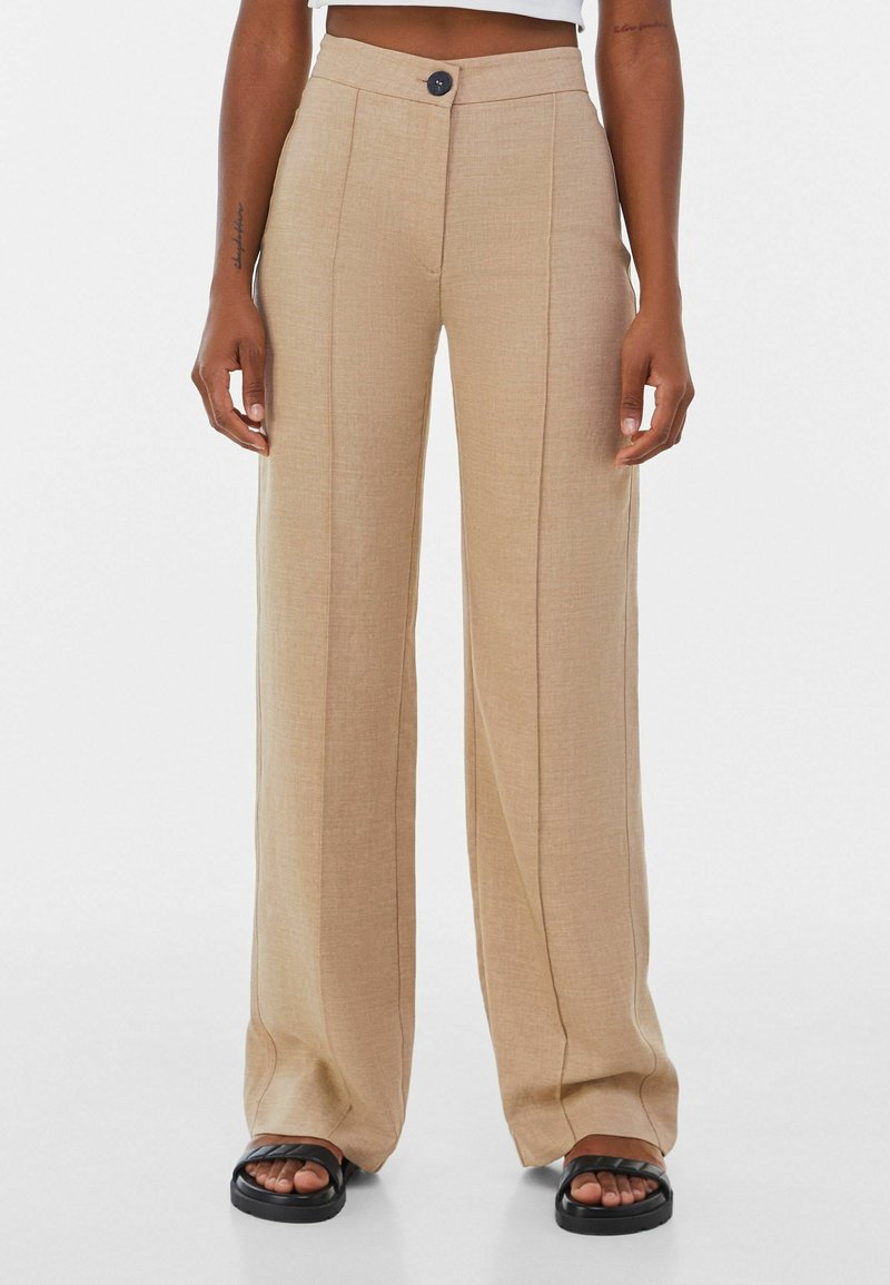 Beige weit geschnittene Hose mit glatter Textur, Bügelfalte und einem einzelnen Knopfverschluss. Tragen mit schwarzen flachen Sandalen.