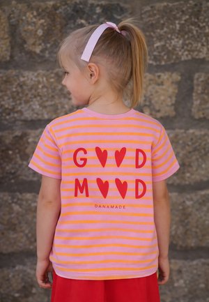 Junges Mädchen mit blondem Haar, das mit einem rosa Band gebunden ist, trägt ein rosa-orange gestreiftes Shirt mit roten Herzen und dem Text "GOOD MOOD" auf dem Rücken.