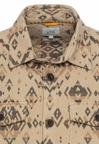 Beige shirt met een geometrisch bruin en zwart patroon. Heeft een puntige kraag, knoopsluiting aan de voorkant en twee borstzakken met bruine knopen.