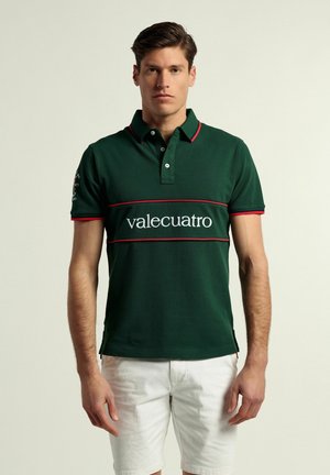 Camisa polo verde de algodón, con cuello, logo blanco "valecuatro" y acentos rojos en el cuello y las mangas. Llevada con pantalones cortos blancos.