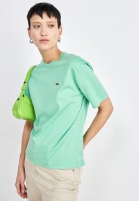 Camiseta de algodón color verde menta con cuello redondo y mangas cortas, que presenta un pequeño logo en el pecho. Llevada con un bolso brillante verde.