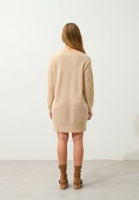 Beige virkad klänning med långa ärmar, ribbat mönster och avslappnad passform. Matchas med bruna strumpor och klackade träskor.