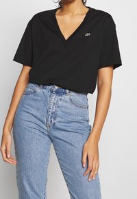 Svart V-ringad T-shirt i bomull med korta ärmar; har en liten grön Lacoste-logotyp på bröstet. Kombinerad med blå högmidjade jeans.