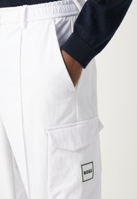 Λευκά cargo παντελόνια με ελαστική μέση, διαθέτοντας πλαϊνή τσέπη και μια μικρή ετικέτα με πράσινο περίγραμμα που γράφει "BOSS". Λείο υλικό.