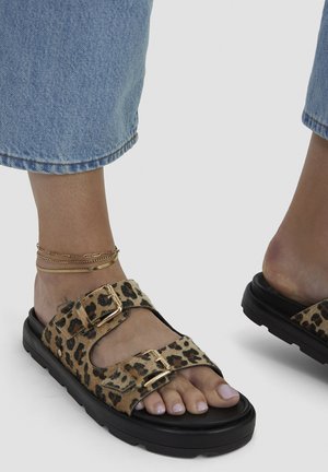 Feet wearing leopard print sandals with double gold buckles, blue denim cropped pants, and a gold layered ankle bracelet.