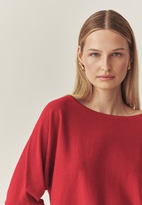 Pull en maille rouge avec un large décolleté et une coupe ample. Le modèle a de longs cheveux lisses et porte des boucles d'oreilles dorées. Fond uni.