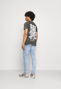 T-shirt gráfica verde desbotada com uma estampa de dragão branco nas costas, combinada com jeans skinny azul claro e ténis dourados prateados.