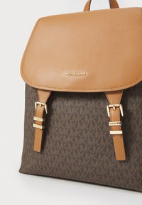 Sac à dos en tissu marron avec rabat en cuir beige, accents dorés et bretelles ajustables. Logo affiché sur le devant. Motif texturé visible.