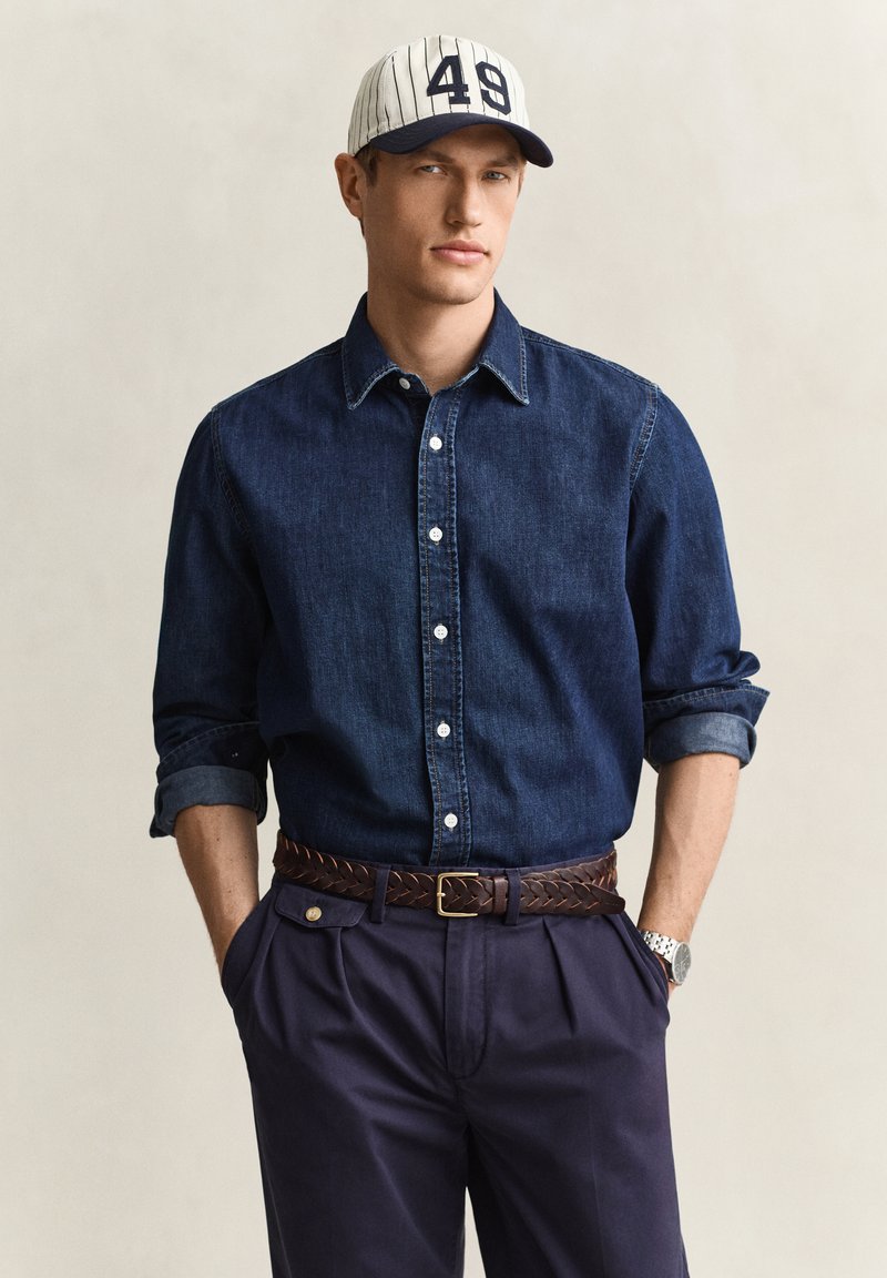 Mann trägt eine weiße und dunkelblaue Baseballkappe, ein dunkles Jeanshemd mit Knöpfen und hochgekrempelten Ärmeln, dunkelblaue Hose und einen geflochtenen braunen Gürtel, Hände in den Taschen.