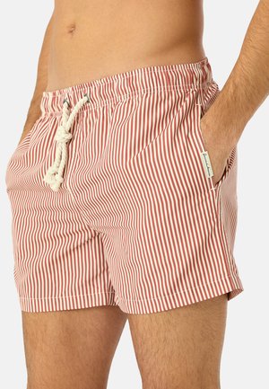 Mann trägt rot-weiß vertikal gestreifte Badeshorts mit weißem Kordelzug, Hand in der Tasche, vor schlichter Hintergrund.